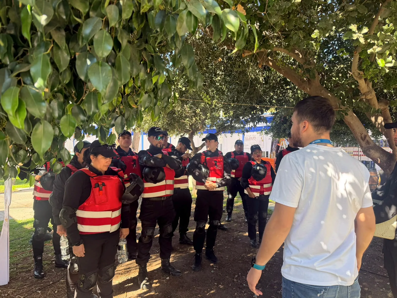 Personal de seguridad con chalecos rojos escuchando indicaciones bajo árboles
